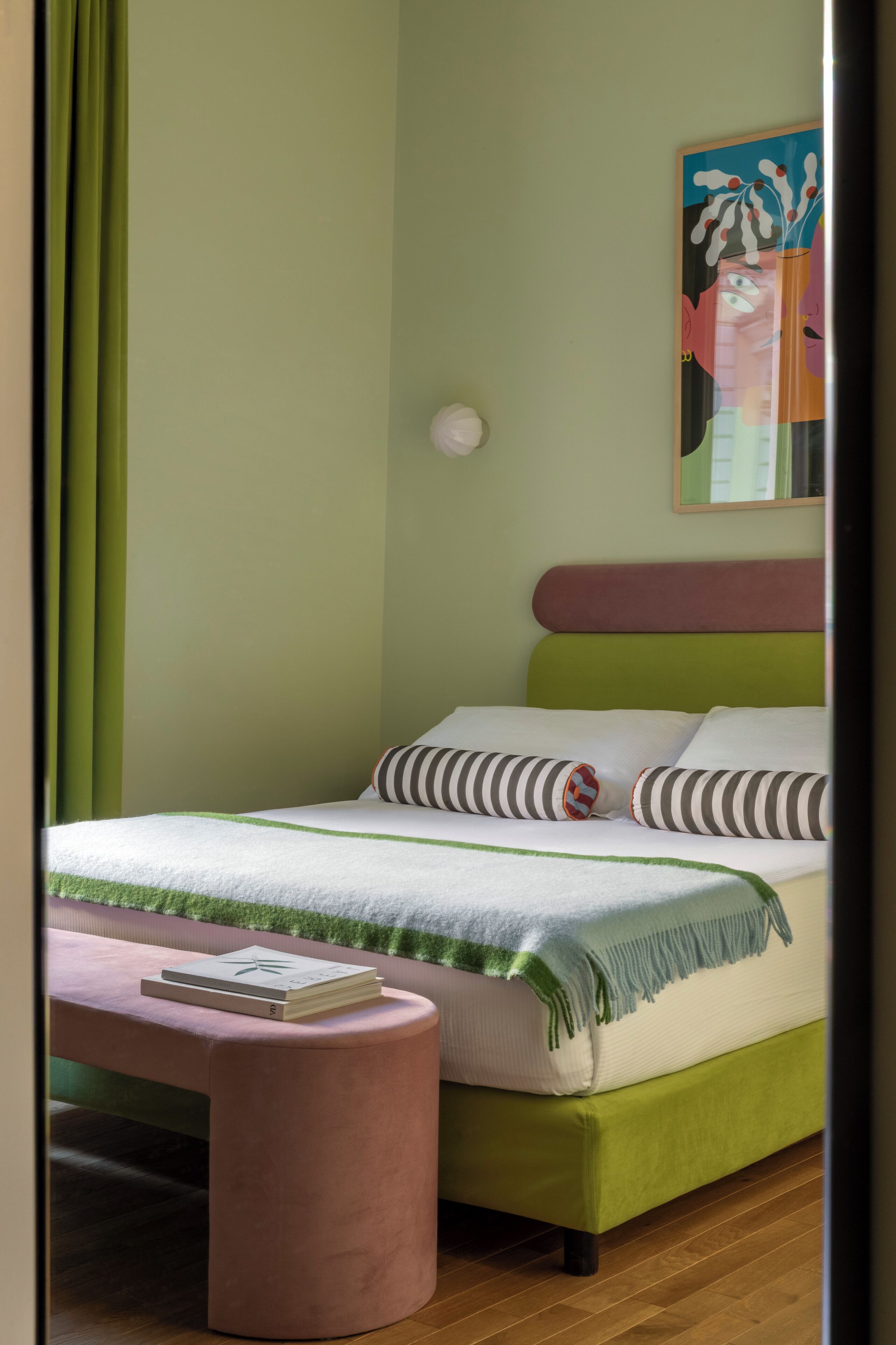 Una cama bien hecha con cabecera verde, un banco rosa con un libro y un cuadro en la pared.