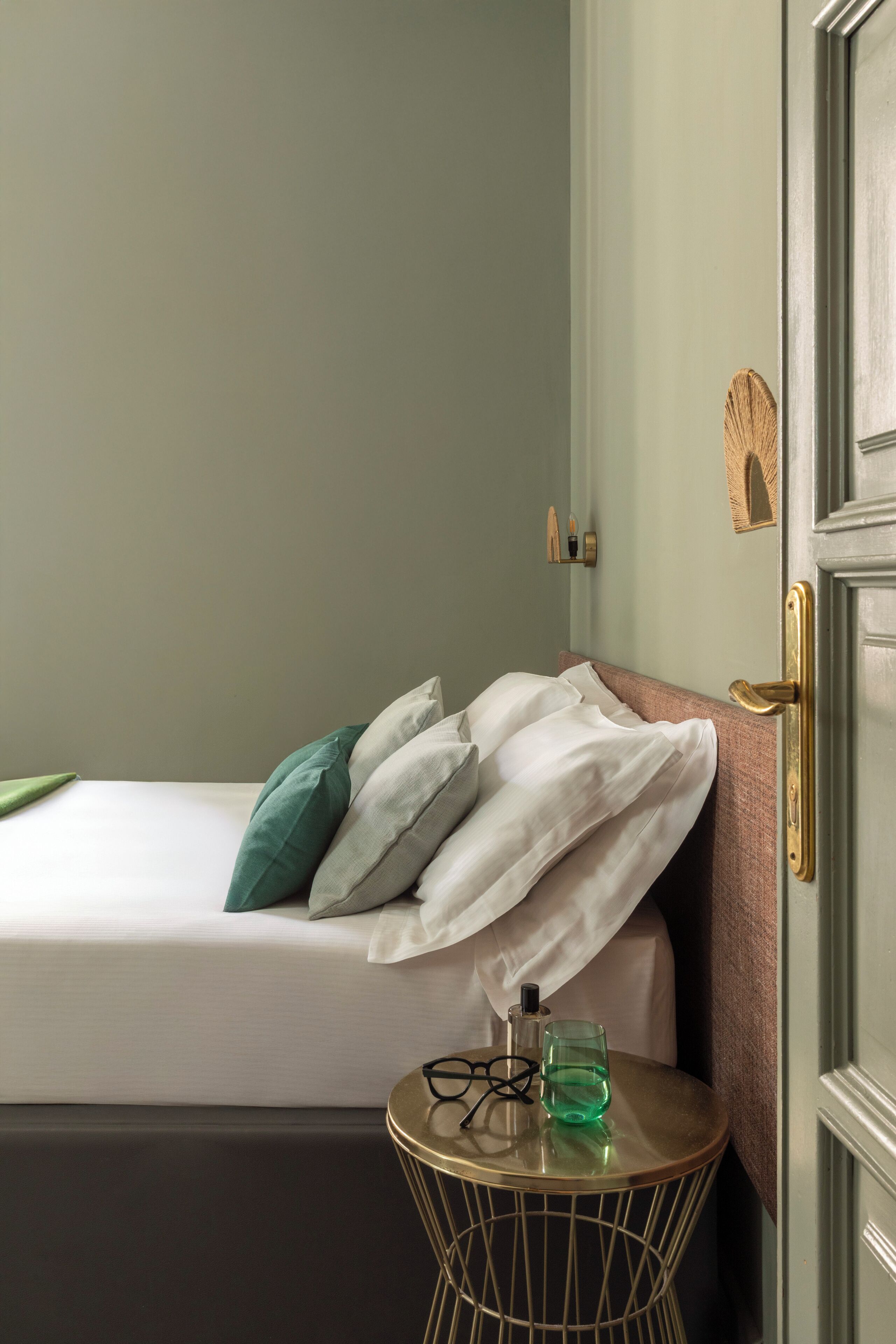 A neatly made bed with pillows, a bedside table with a glass of water and glasses, and a door with a brass handle.