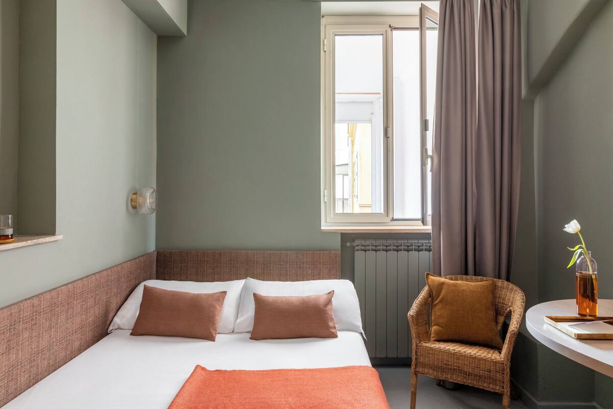 Hotellihuone, jossa on sänky, tuoli, pieni pöytä maljakolla ja ikkuna, jossa on verhot.