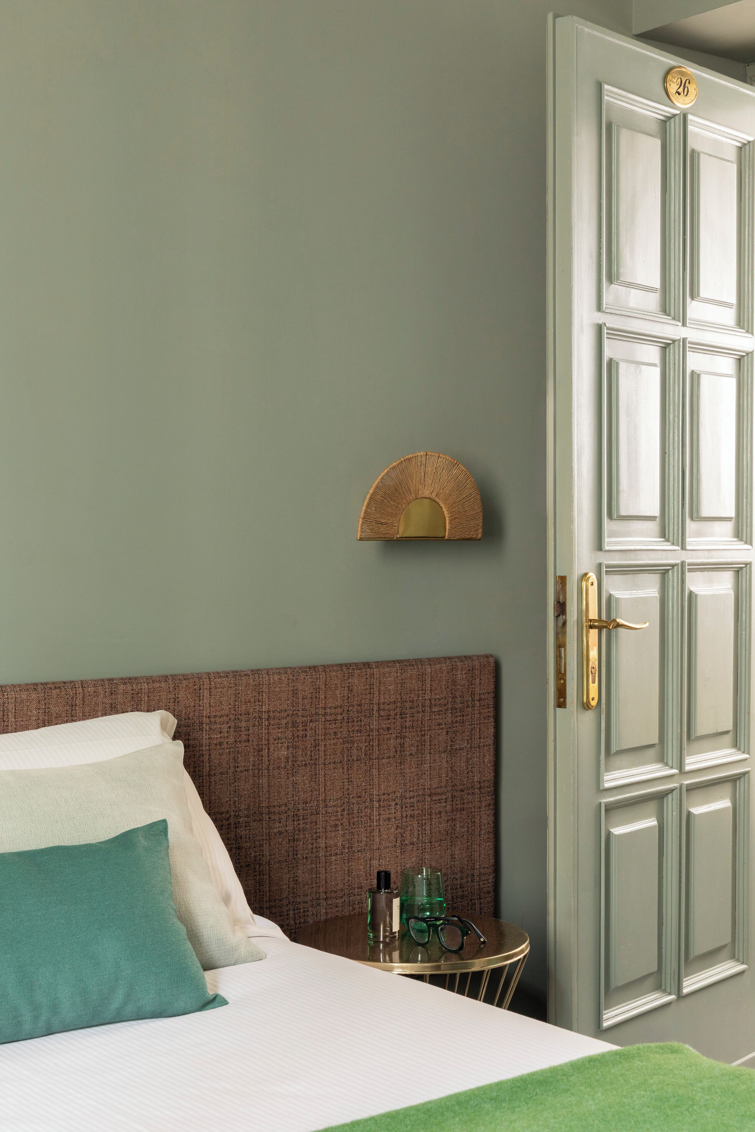 A bedroom with a bed, a bedside table with toiletries, a wall-mounted lamp, and a door with a number plate.