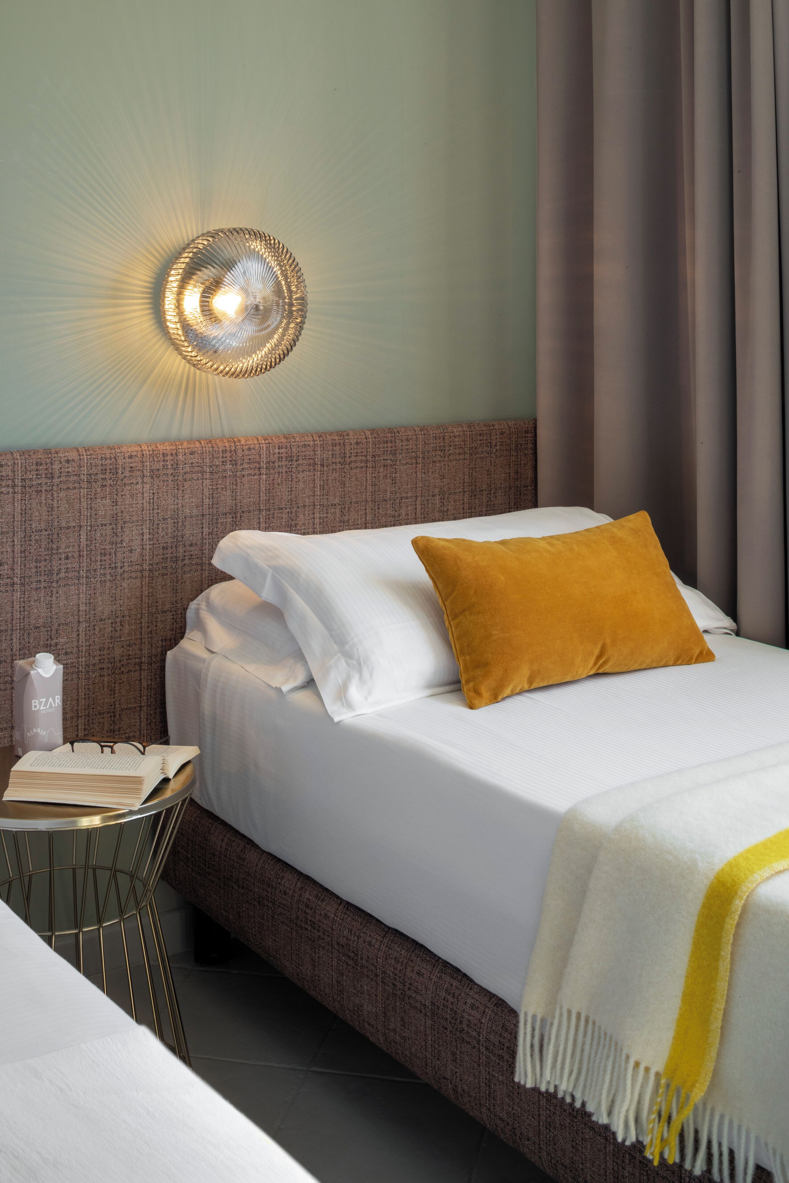 Cama bem arrumada com lençóis brancos e um travesseiro amarelo-mostarda, mesa de cabeceira com um livro e uma garrafa, e luminária fixada na parede.