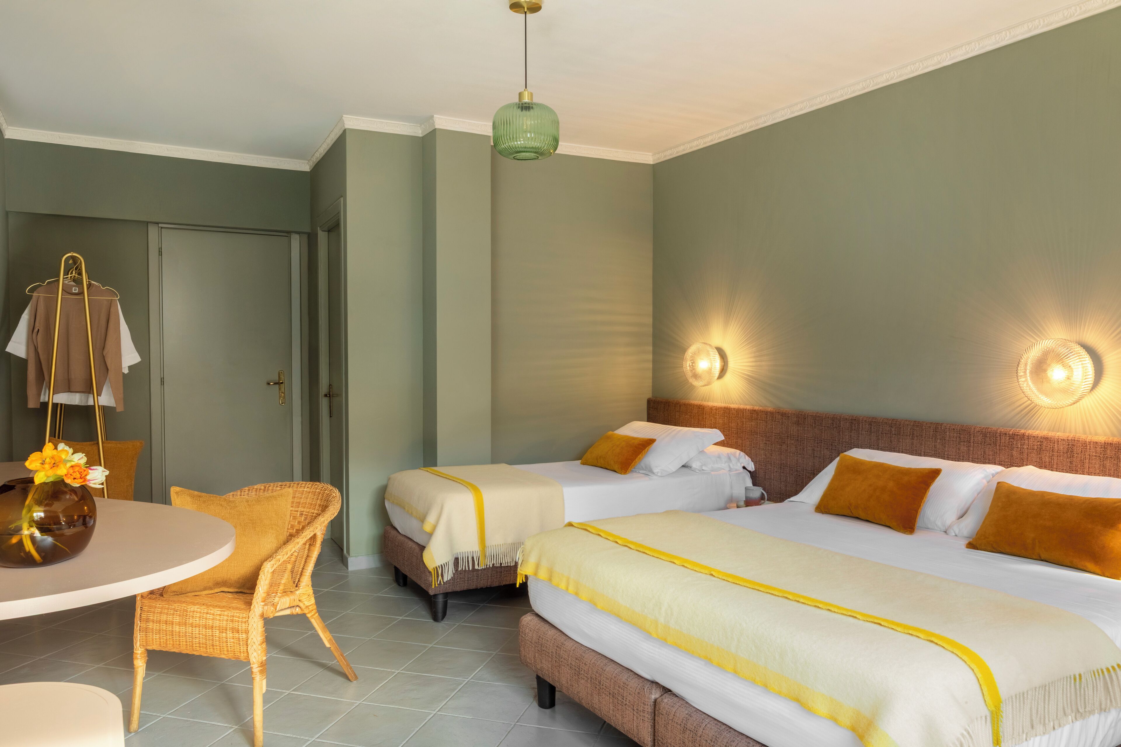 Une chambre d’hôtel avec deux lits, une table à manger avec des chaises, un fauteuil suspendu et un vase avec des fleurs.