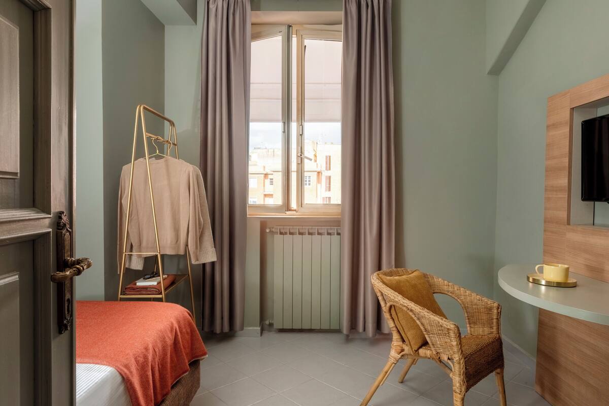 Hotellihuone, jossa on sänky, tuoli, pieni pöytä, jossa on kuppi, ikkuna verhoilla ja patteri.