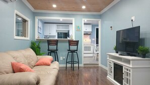 Un salon avec un canapé beige, deux fauteuils, un téléviseur et une cuisine en arrière-plan.