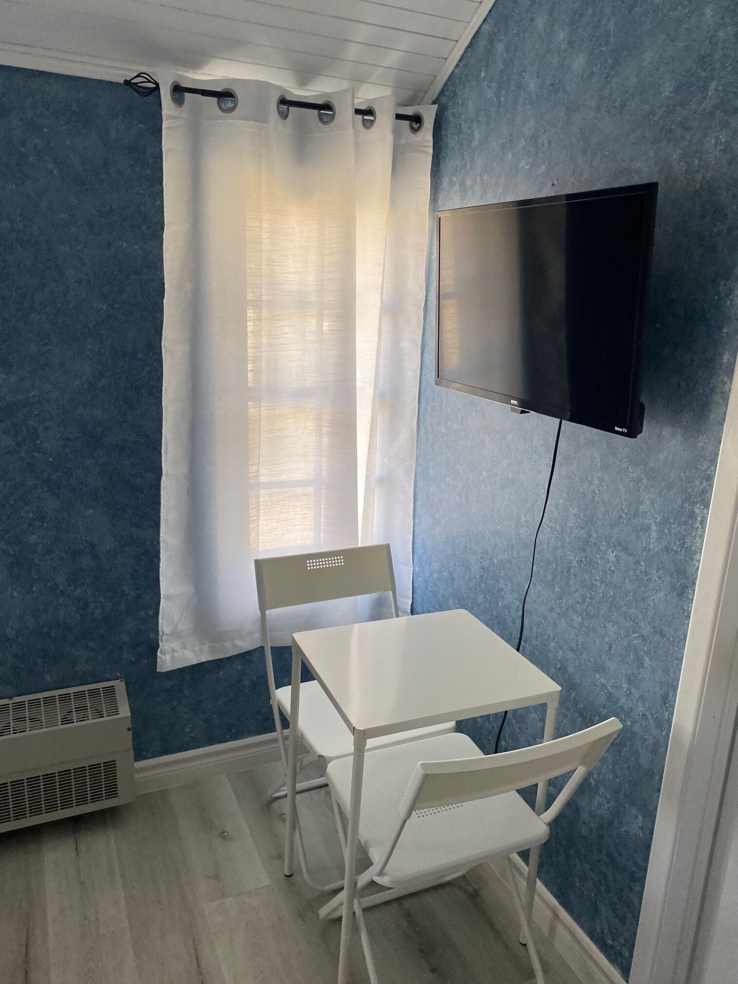 Una habitación pequeña con una mesa blanca y dos sillas, un televisor montado en la pared, una ventana con cortina blanca y un respiradero en el suelo.