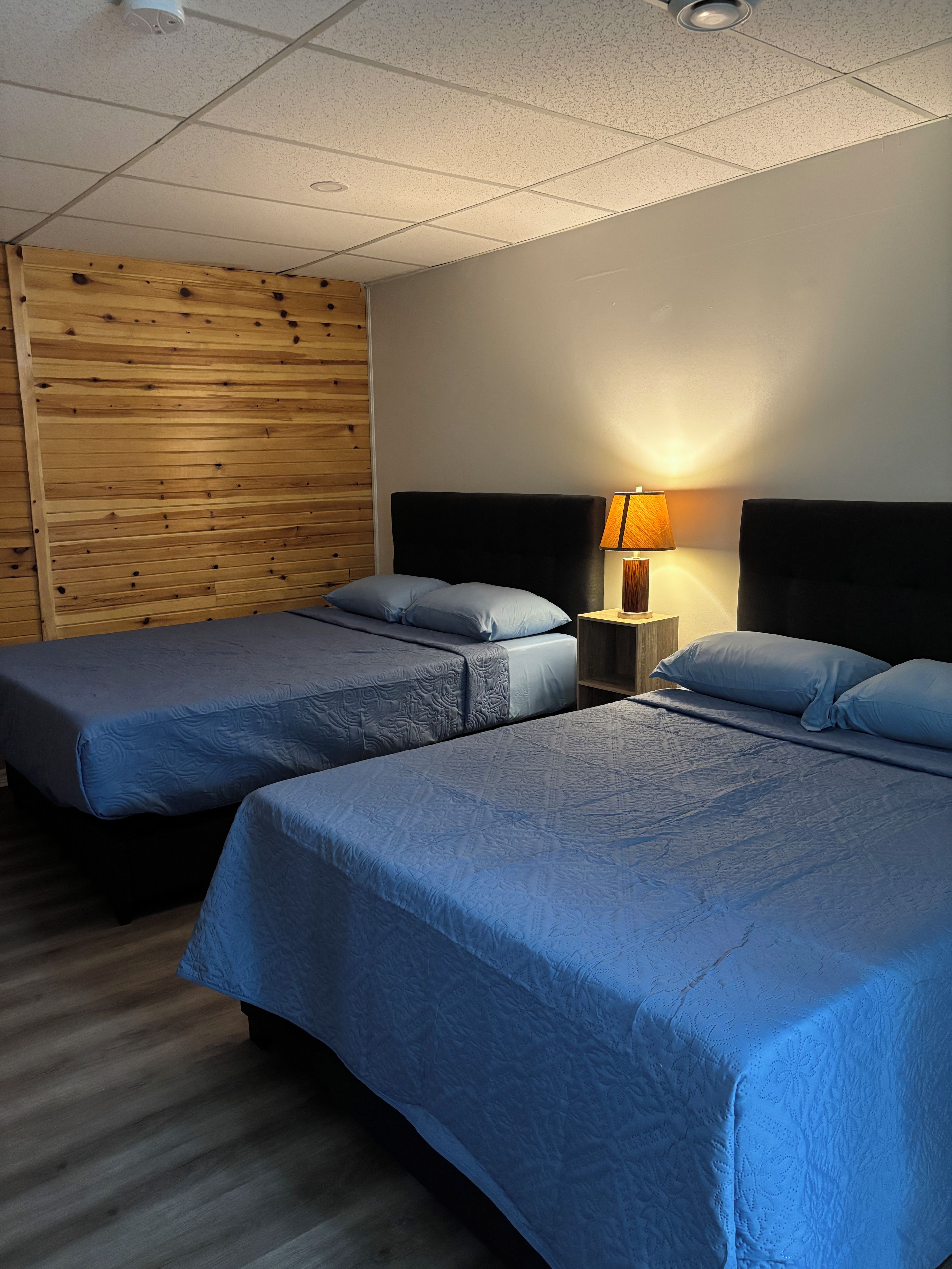 A hotel room with two beds, a wooden panel wall, a nightstand with a lamp, and a ceiling with recessed lighting.
