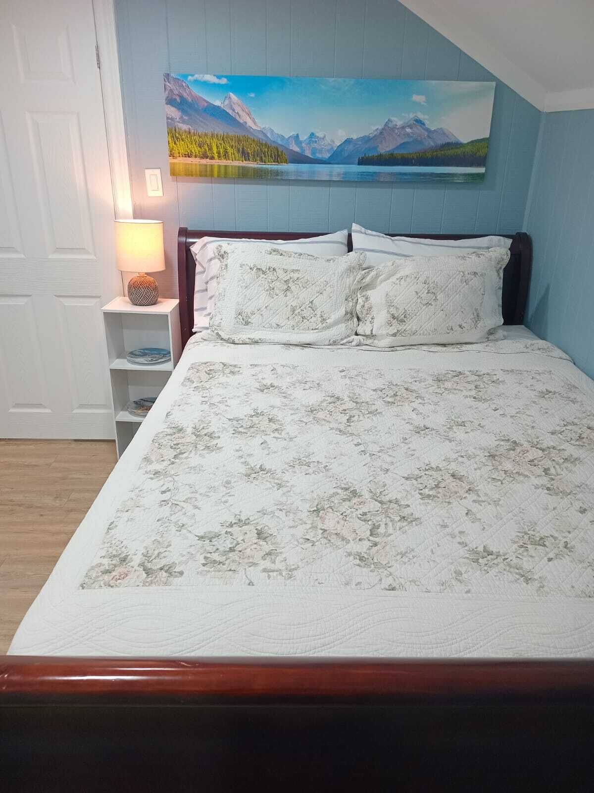 Een slaapkamer met een bed, nachtkastje, lamp en een schilderij van een berglandschap.