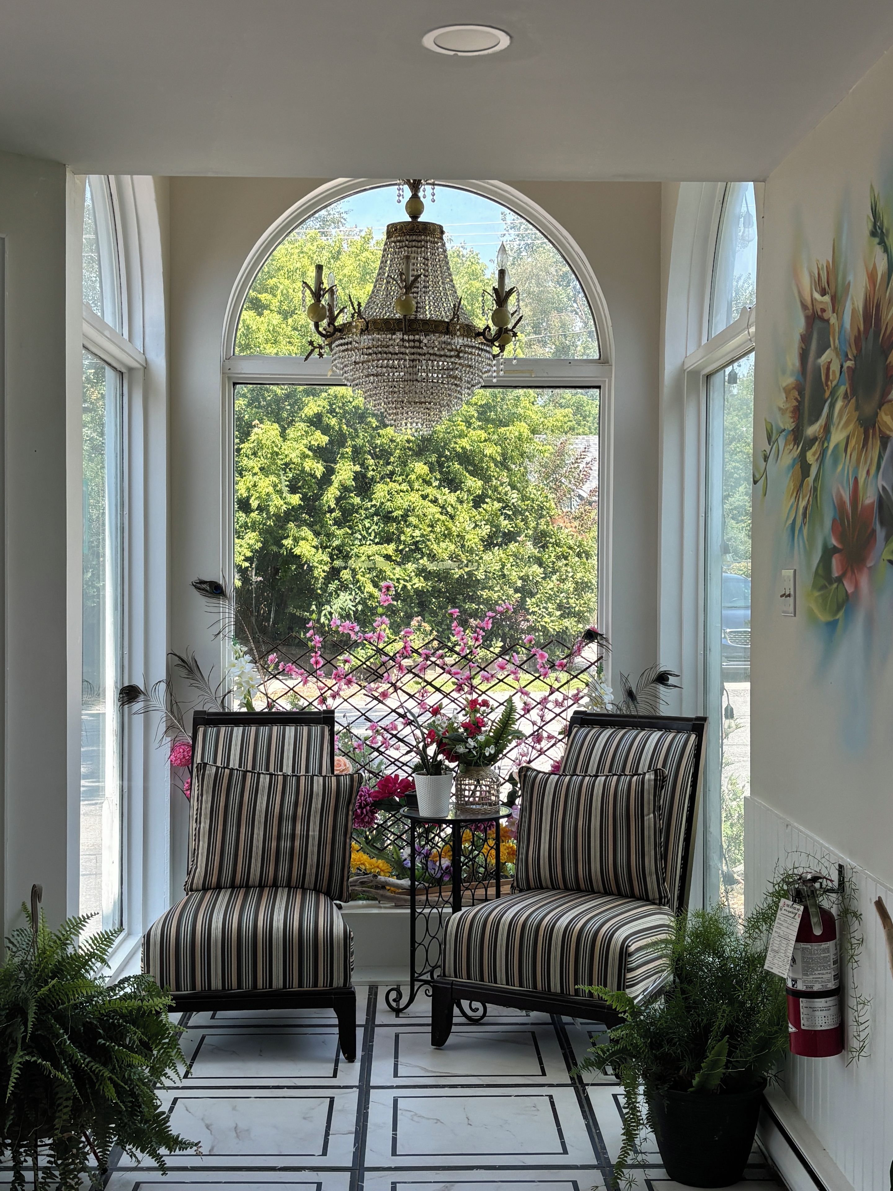 A room with two striped armchairs facing a large arched window, a chandelier, and a fire extinguisher on the wall.
