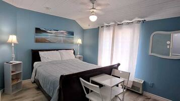 A bedroom with a bed, a desk, and a chair. There is a window with curtains, a ceiling fan, and a painting on the wall.
