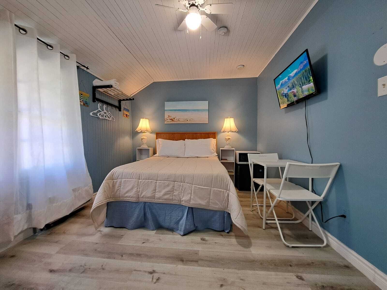 Un dormitorio con cama, dos mesitas de noche con lámparas, un televisor en la pared y un escritorio pequeño con sillas.