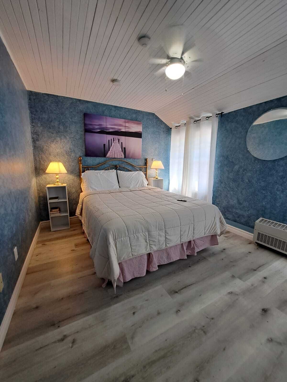 Een slaapkamer met een bed, twee nachtkastjes met lampen, een plafondventilator, een wandairconditioner en een groot raam met gordijnen.