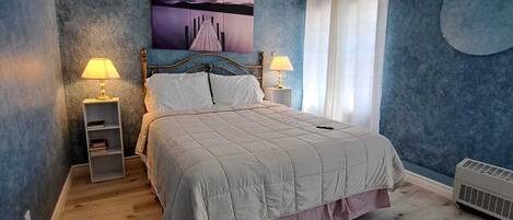 A bedroom with a bed, two bedside tables with lamps, a ceiling fan, a wall-mounted air conditioner, and a large window with curtains.