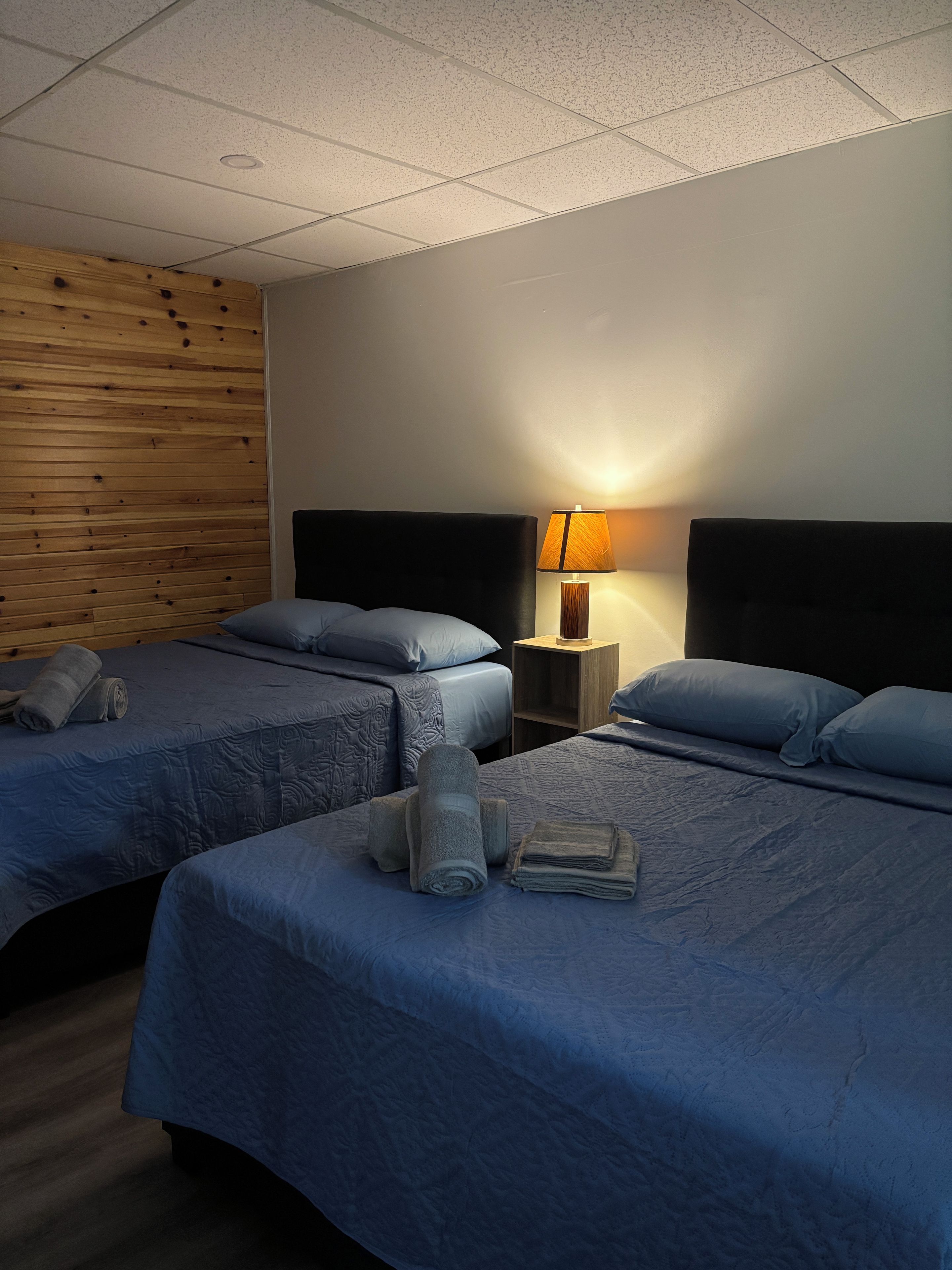 A hotel room with two beds, a wooden accent wall, a nightstand with a lamp, and a neatly made bed with towels.