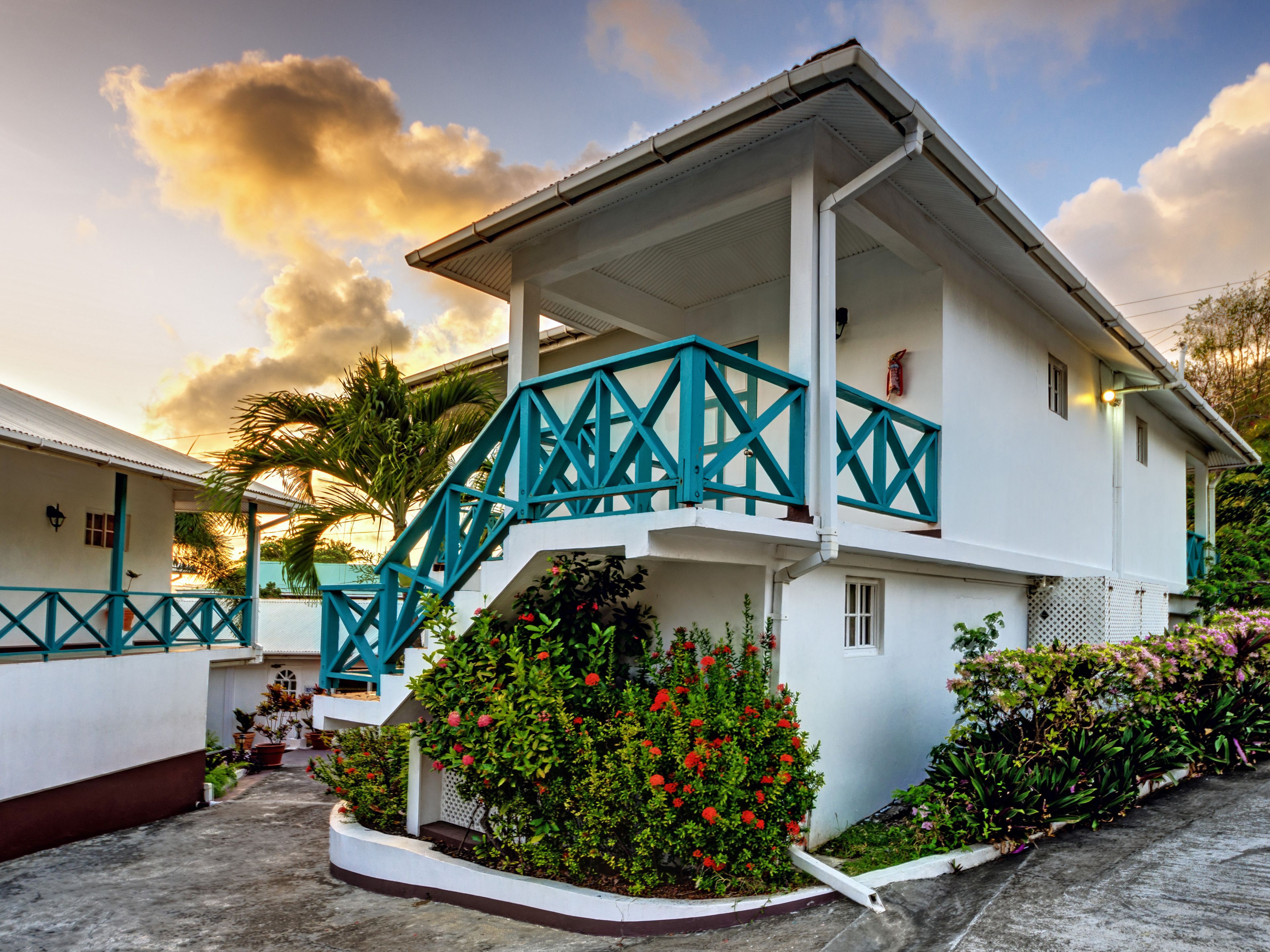 A white building with a blue staircase and balcony, surrounded by lush greenery and flowering plants.