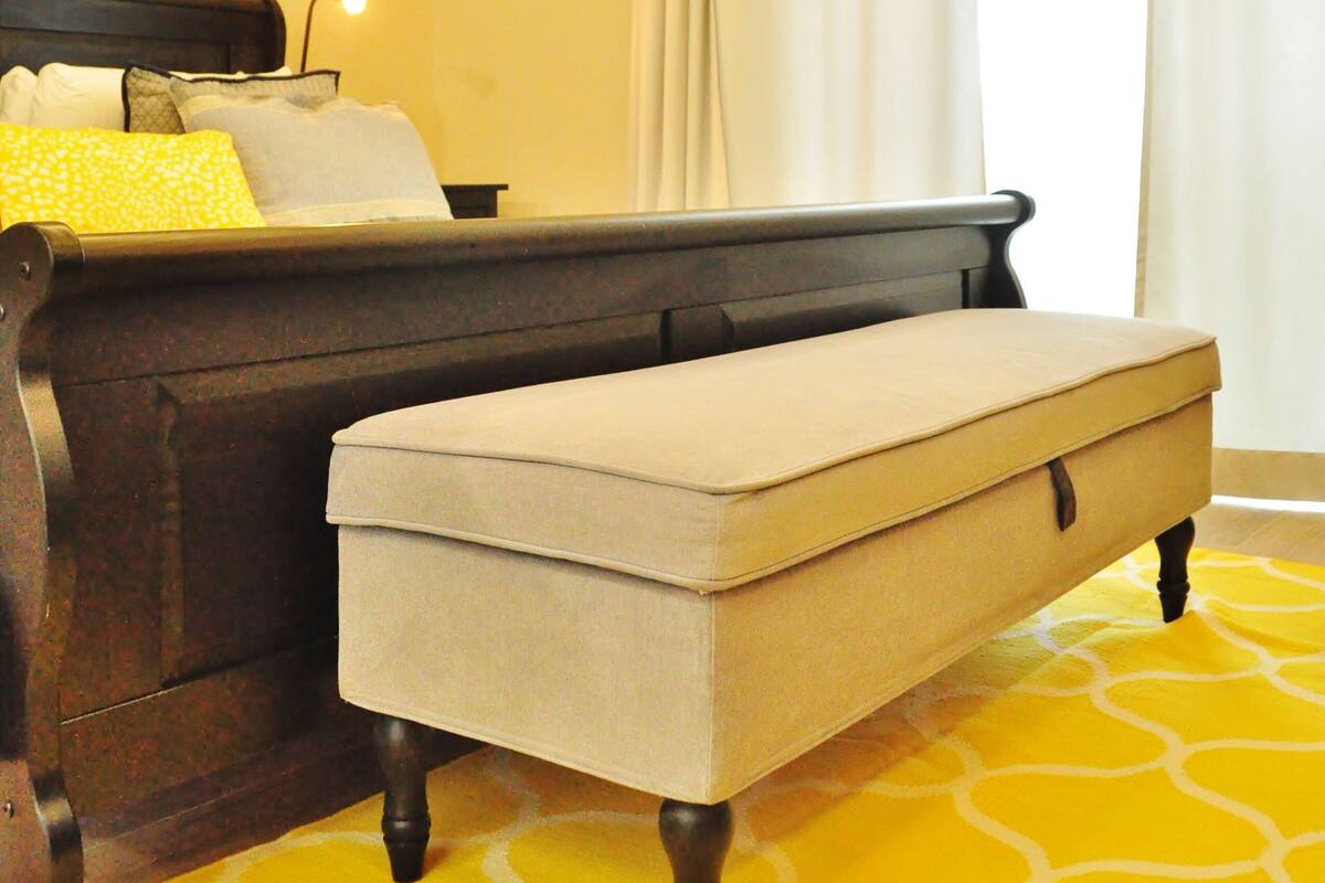 Une pièce comprenant un lit, un banc, un tableau encadré au mur et une fenêtre avec des rideaux.
