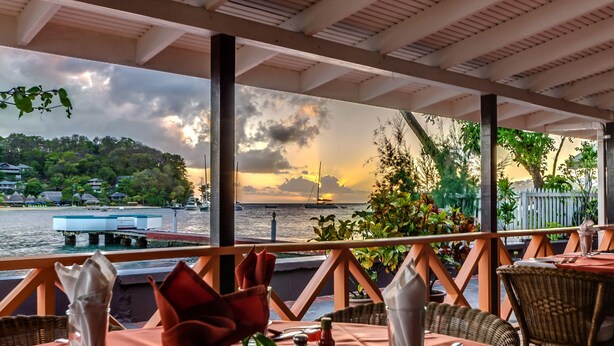 An outdoor dining area with tables set for two, overlooking a body of water with a sailboat and a dock. - Mariners Hotel (Villa)