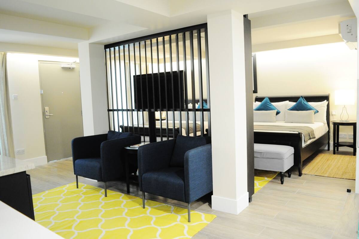 Une chambre d’hôtel moderne avec un lit, deux fauteuils, une télévision et un tapis jaune à motifs.