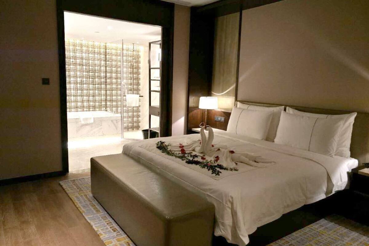 Hotelkamer met een groot bed, een bankje aan het voeteneind, een nachtkastje met lamp en uitzicht op de badkamer door een open deur.