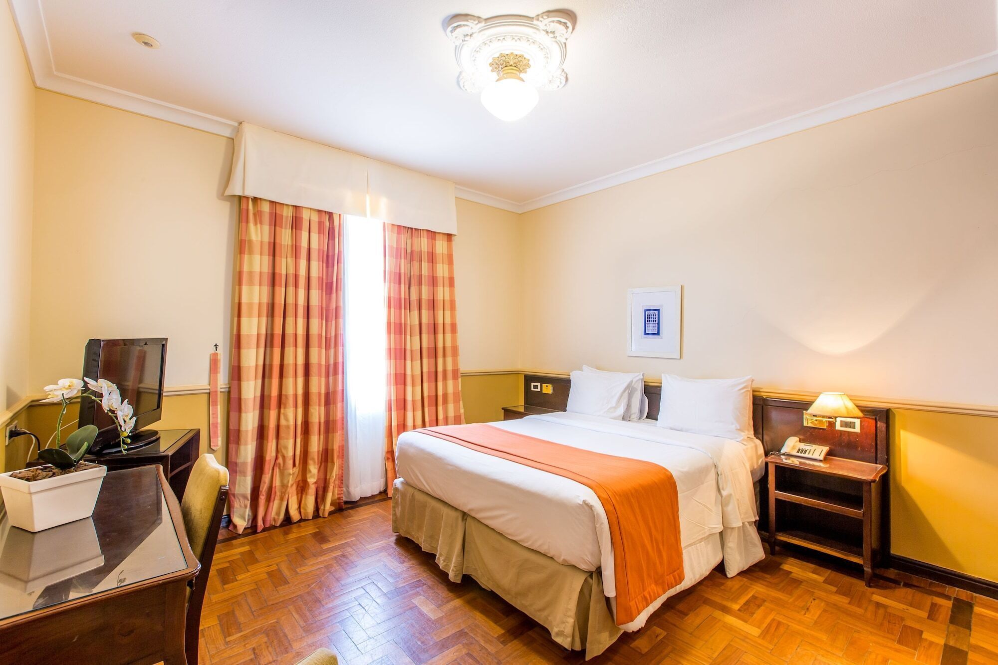 Quarto de hotel com cama, mesa de cabeceira, abajur e escrivaninha com planta.