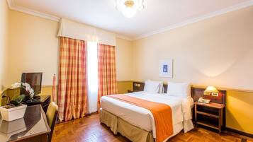 Quarto de hotel com cama, mesa de cabeceira, abajur e escrivaninha com planta.