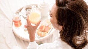 Una persona sentada en una cama con una bandeja de desayuno que incluye tostadas, un vaso de jugo de naranja y una tetera.