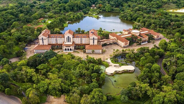 Vista aérea de un edificio grande e histórico con varias secciones, rodeado de abundante vegetación y un lago central.