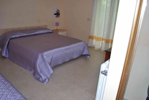 A bedroom with a large bed, a nightstand, a lamp, and a window with curtains. - Villa Archegeta (Giardini Naxos)
