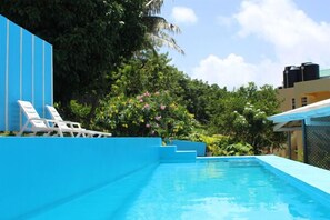 A swimming pool with steps leading into it, surrounded by greenery and a blue building. - Hillside Apartments (Villa)