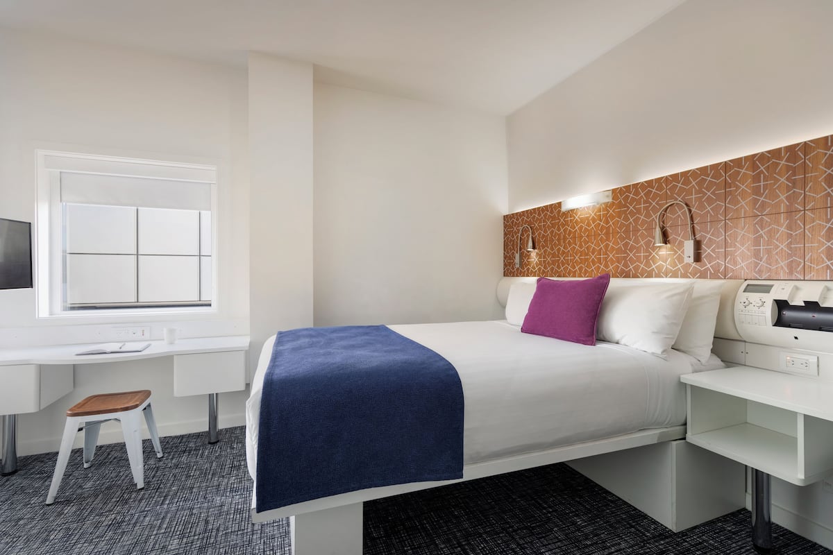 Habitación de hotel con cama, escritorio, silla, televisor y panel de control en la pared.