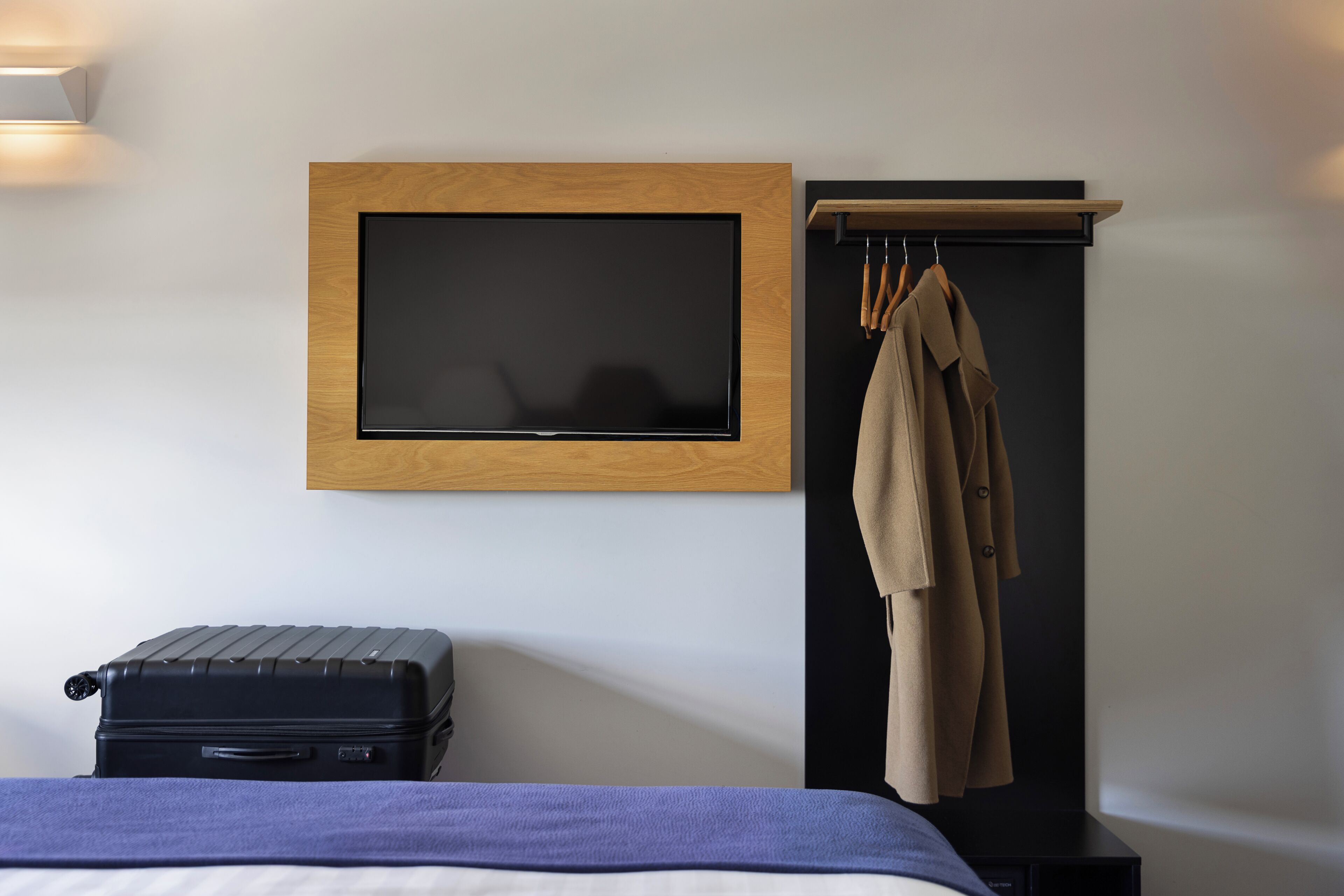 A hotel room with a bed, a flat-screen TV mounted on the wall, a coat rack with a coat, and a suitcase on the floor.