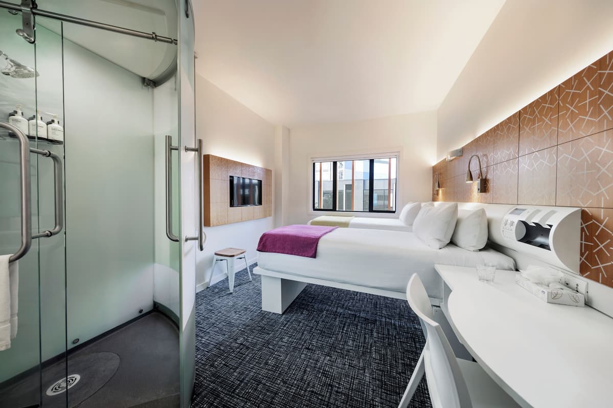 Habitación de hotel con cabina de ducha de vidrio, cama con ropa blanca, una mesita pequeña y una zona de estar con sofá blanco.