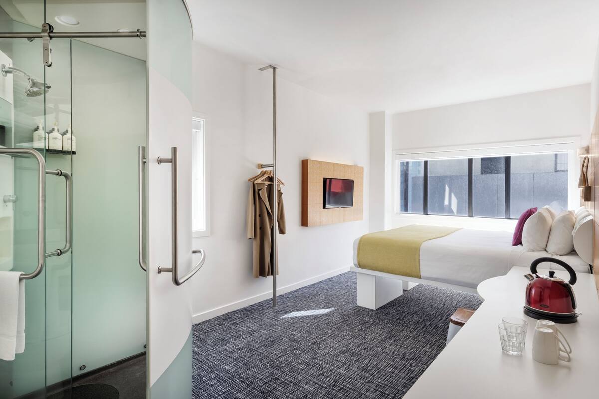 Una habitación de hotel moderna con cabina de ducha de vidrio, una cama con cabecera blanca, una mesita de noche con hervidor y una ventana con vistas a la ciudad.
