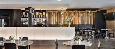 A modern bar area with a counter, bar stools, and a variety of bottles. Tables with chairs are arranged in the dining area.