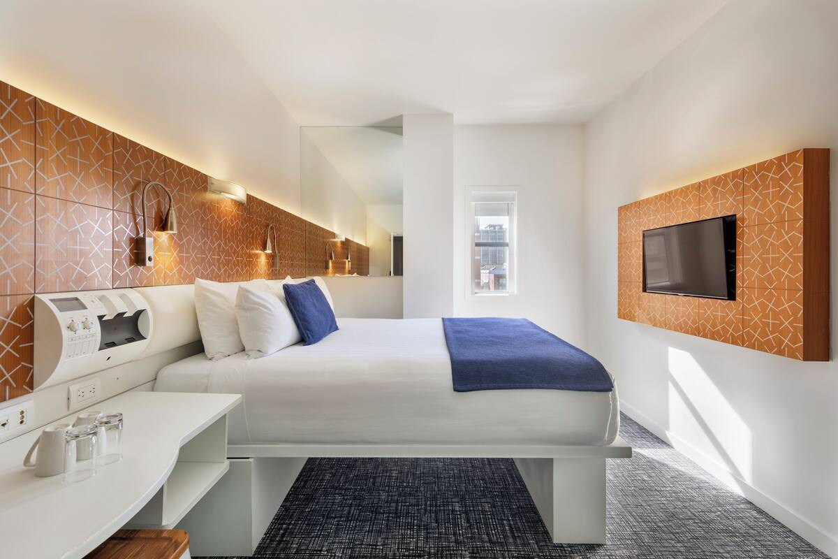 Habitación de hotel con cama, mesita de noche, televisión y panel de control en la pared.