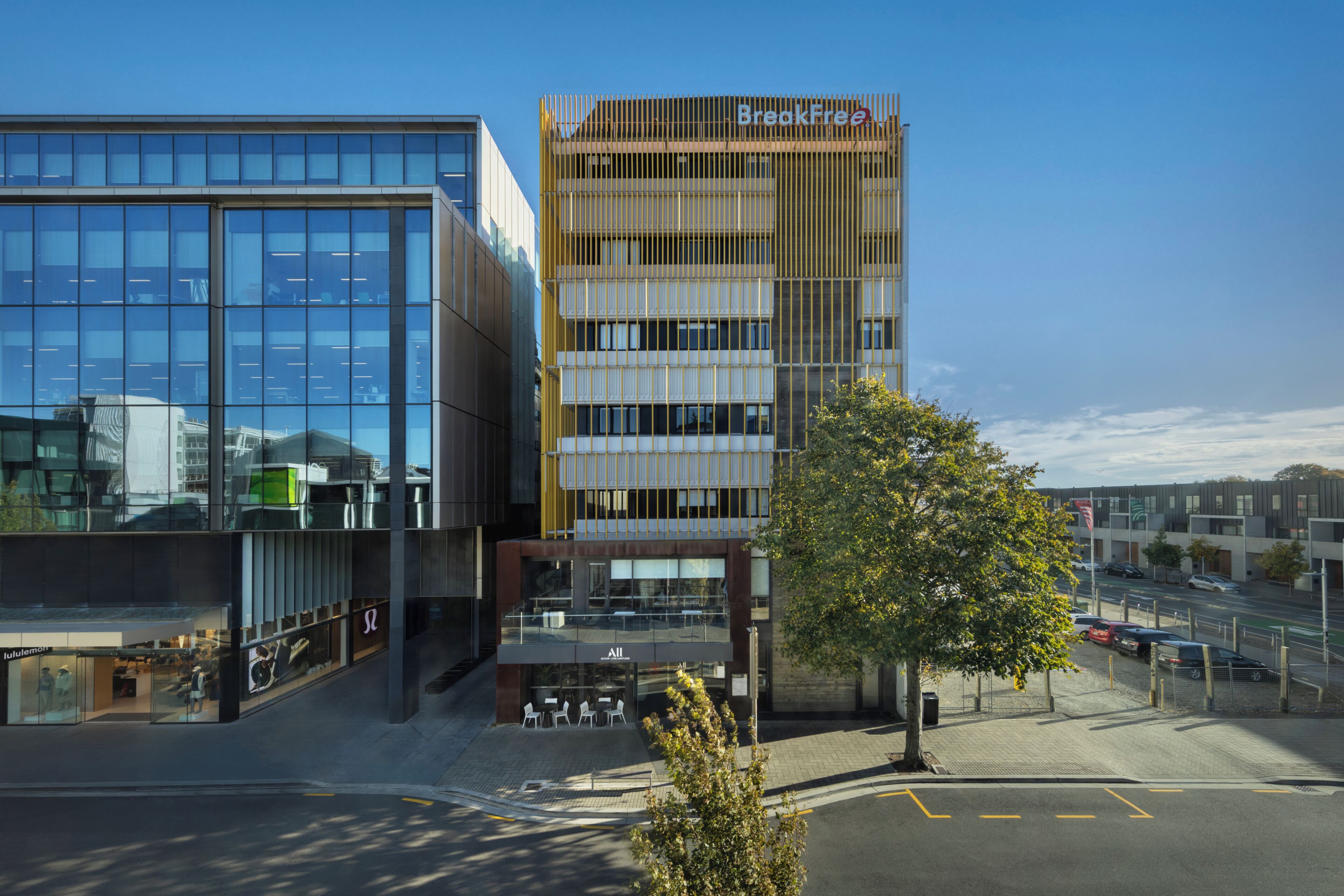 A modern building with a glass facade, a wooden panel design on the upper floors, and a tree in the foreground.