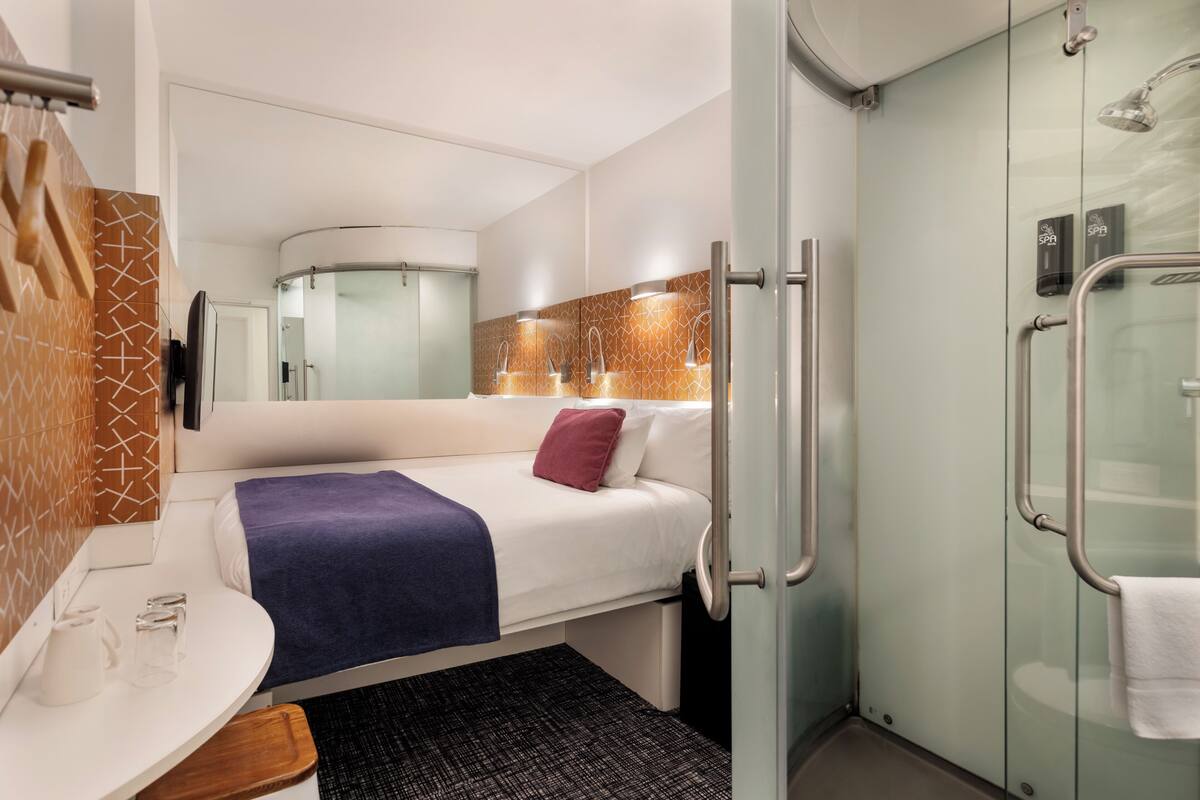 Una habitación de hotel moderna con una cama, una mesita con hervidor, zona de ducha y baño con mampara de vidrio.