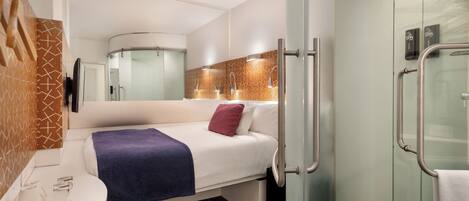 Quarto de hotel moderno com cama, uma mesinha com chaleira, área de chuveiro e banheiro com divisória de vidro.