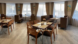A hotel dining area with multiple tables and chairs, large windows with curtains, and artwork on the walls.