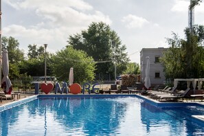 Une piscine entourée d’arbres et d’un bâtiment, avec des chaises longues et des parasols.