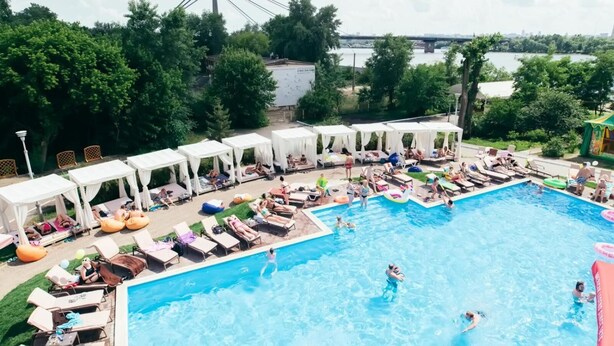 A swimming pool with lounge chairs and umbrellas, surrounded by trees and a bridge in the background.
