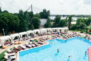 A swimming pool with lounge chairs and umbrellas, surrounded by trees and a bridge in the background. - Vita Park Borysfen (Kyiv)