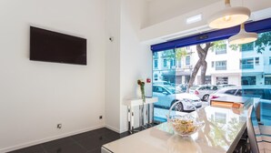 A modern hotel room with a large window, a flat-screen TV mounted on the wall, a dining table with a vase of flowers, and a view of parked cars outside. - Hotel Capital (Lisbon)