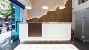 A modern reception desk with a wooden accent wall, pendant lighting, and a glass door leading outside. - Hotel Capital (Lisbon)
