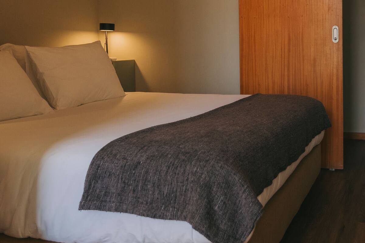 A neatly made bed with a gray blanket, a bedside lamp, and a wooden wardrobe in a hotel room.