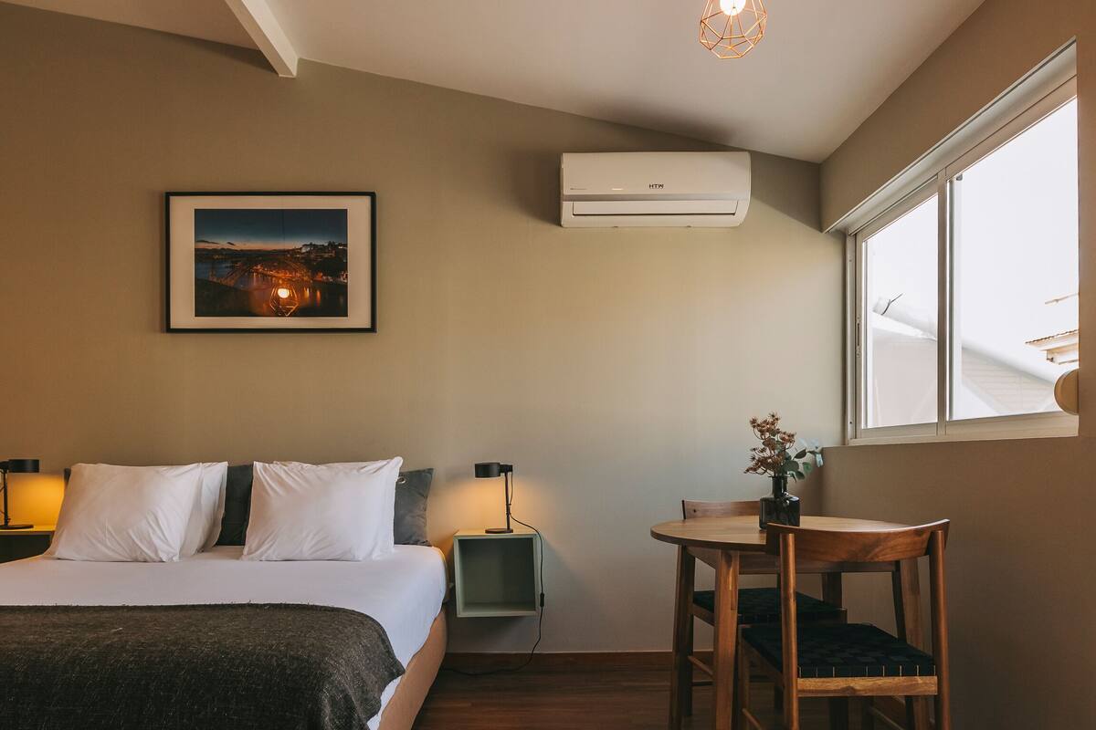 A bedroom with a bed, bedside tables, a desk, and a chair. There is a window, an air conditioner, and a framed picture on the wall.