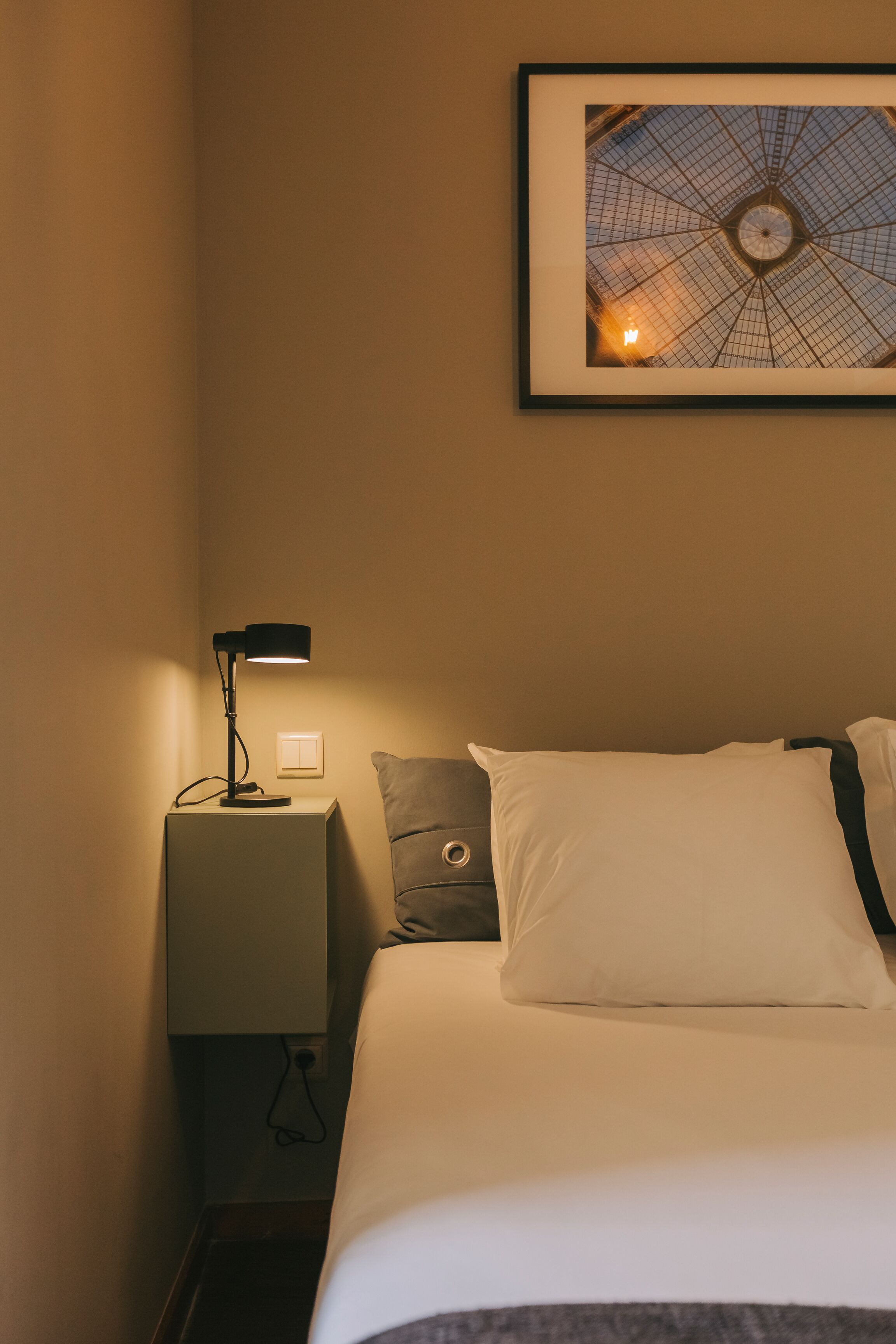 Een net opgemaakt bed met witte lakens, een grijs kussen en een zwarte lamp op een nachtkastje. Een ingelijst beeld hangt aan de muur boven het bed.