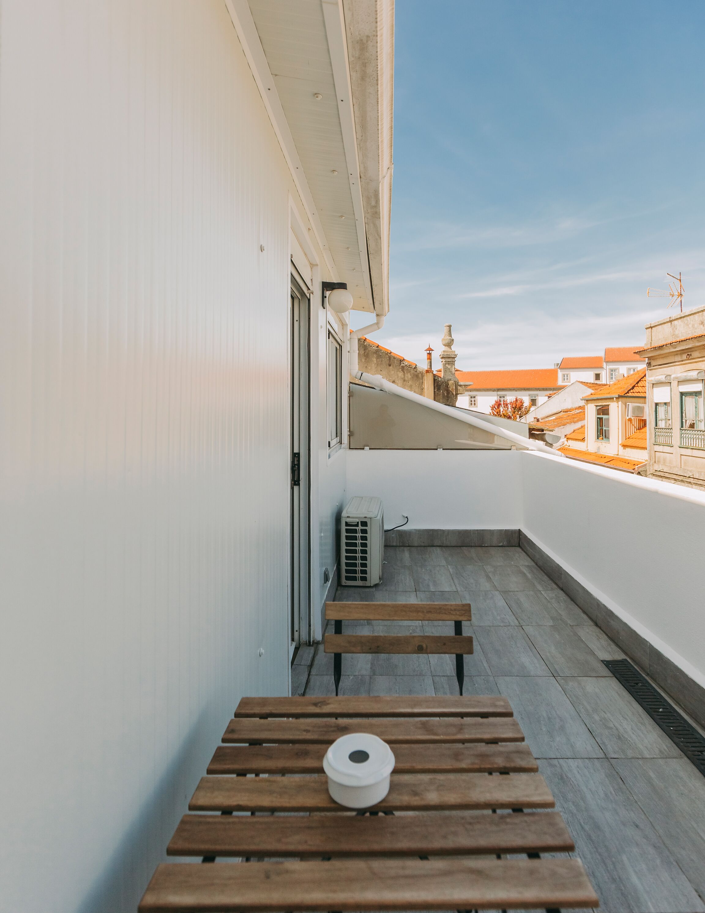 A balcony with a wooden table and a white object on it, a white wall, and a view of buildings with orange roofs.