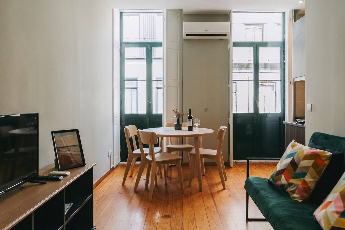 A modern living room with a wooden floor, a green sofa, a dining table with chairs, a TV, and a pendant light.