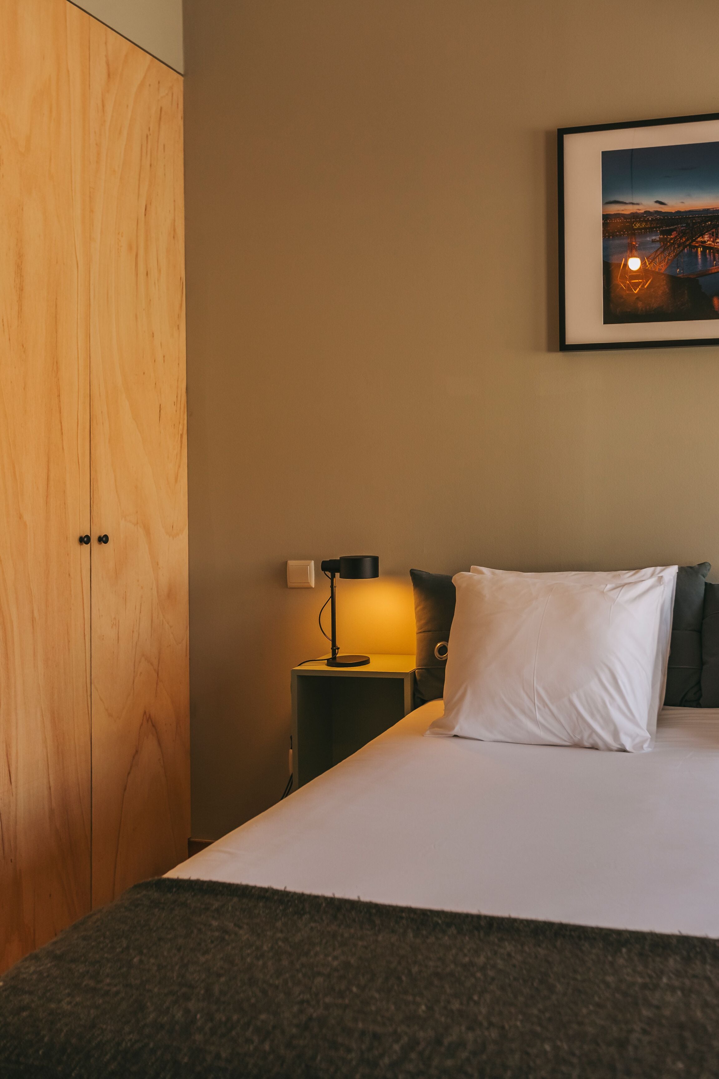 Et hotelværelse med en seng, et natbord med lampe, et billede i ramme på væggen og en træskab.