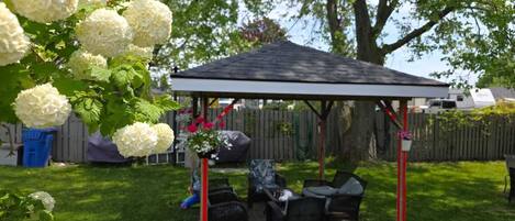 A covered patio area with outdoor furniture, a grill, and a garden with white flowers.